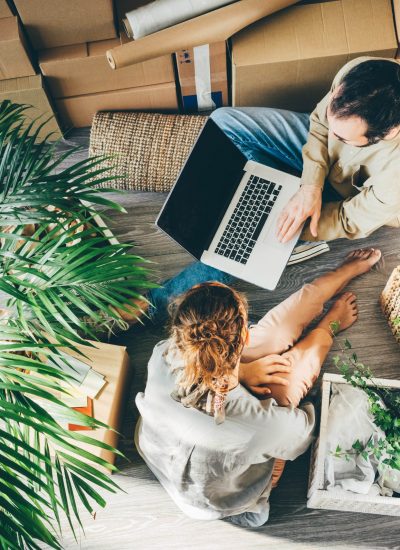 cheerful young family of man and woman wearing casual clothes makes project of decoration for new apartment on laptop sitting on floor near pile of boxes